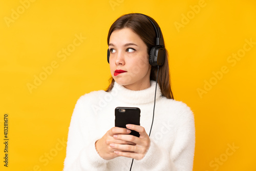 Teenager girl isolated on yellow background listening music with a mobile and thinking