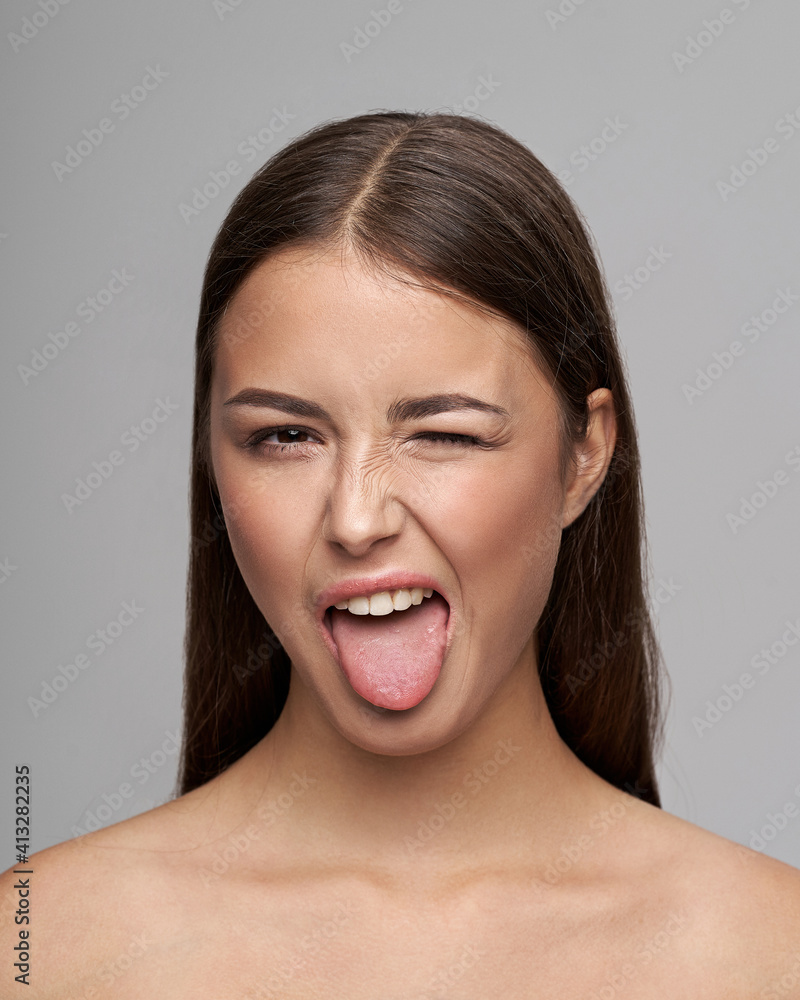 Young beautiful female model close-up studio beauty portrait. Girl with natural make-up and long brunette straight hair. Happy crazy woman shoing her tongue and winking Young beautiful female model close-up studio beauty portrait. Girl with natural make-up and long brunette straight hair. Happy crazy woman shoing her tongue and winking