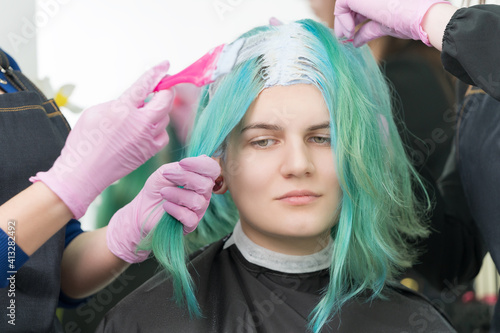 Process of bleaching hair roots in hair salon. Two hairdressers in protective glove use pink brush while applying paint to young adult Caucasian customer with long emerald hair color