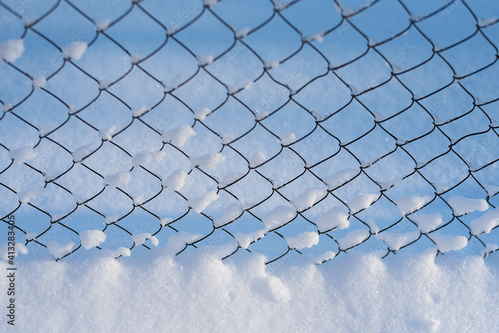 A snow-covered black mesh fence is positioned frontally in the frame. Sunny frosty day in the city. Blurred background. Selective focus. Close plan. Background. A snow-covered black mesh fence is positioned frontally in the frame. Sunny frosty day in the city. Blurred background. Selective focus. Close plan. Background.