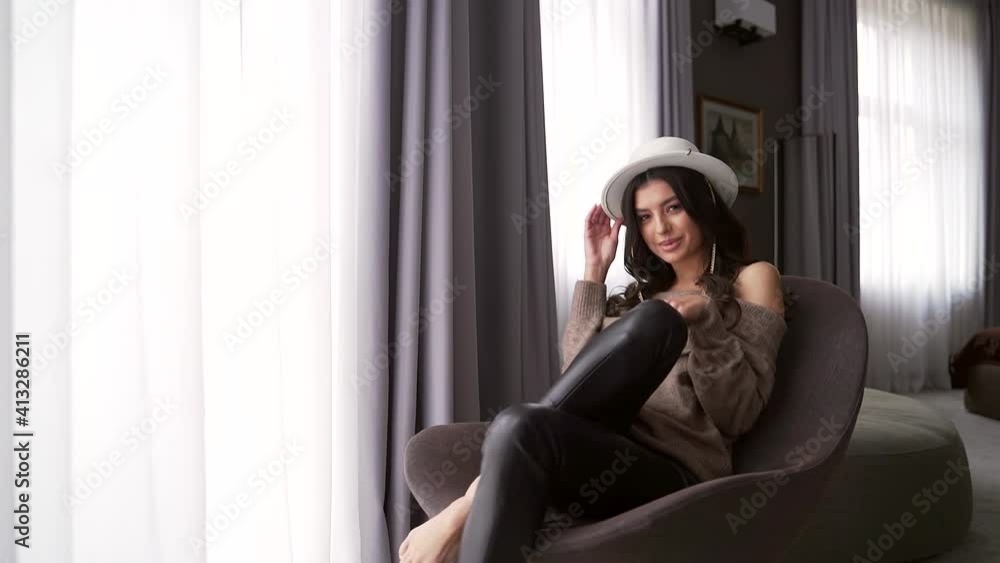 portrait of a young charming and sexy woman and sitting in a chair crossed legs indoors hotel or apartment. Attractive fashion lady, model in a hat looks seductively into the camera.