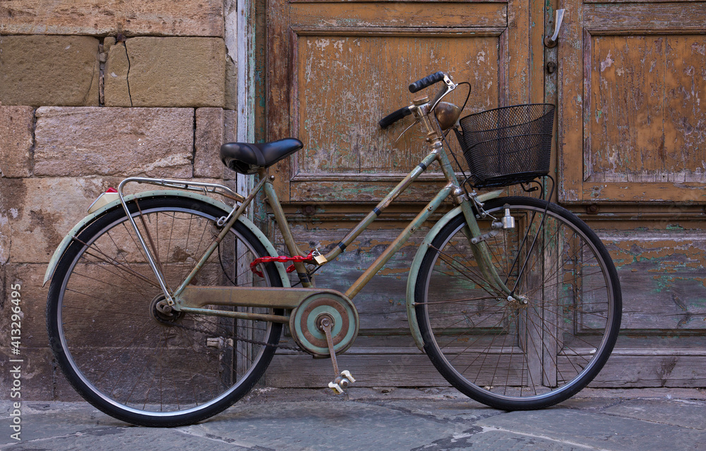 An classic, old, vintage bicycle with wicker basket rests against a weathered wall and door in Lucca, Italy

