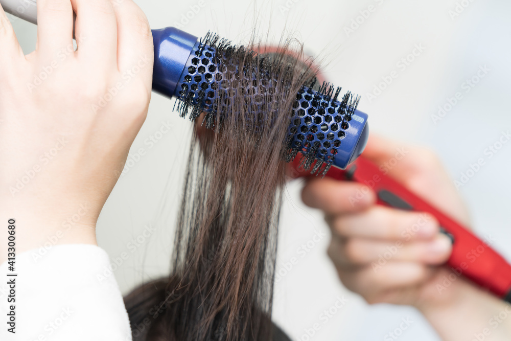 Hands of hairdresser dries brunette hair of client using blue hairbrush and red hairdryer in professional beauty salon. Hands of hairdresser dries brunette hair of client using blue hairbrush and red hairdryer in professional beauty salon.