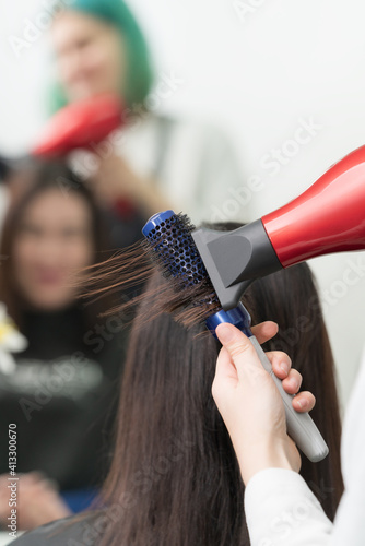 Hands of hairstylist dries brunette hair of client using red hair dryer and blue comb in professional beauty salon.