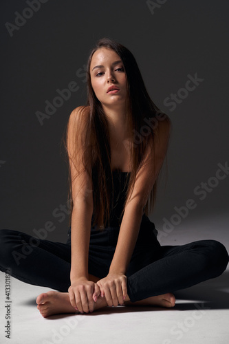 Full length studio portrait of young slim tanned caucasian girl in black jeans and bando top sitting and posing against grey studio background © Dmitry Tsvetkov
