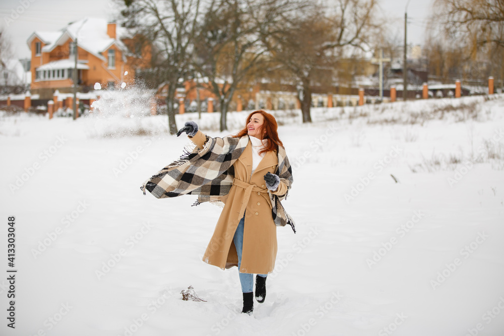 Fototapeta premium Happy woman in winter snow park wearing beige coat and large plaid scarf. Romantic walk in rustic style. Female in casual classic clothes with red hairs. Redhead smiling young girl with dried flowers