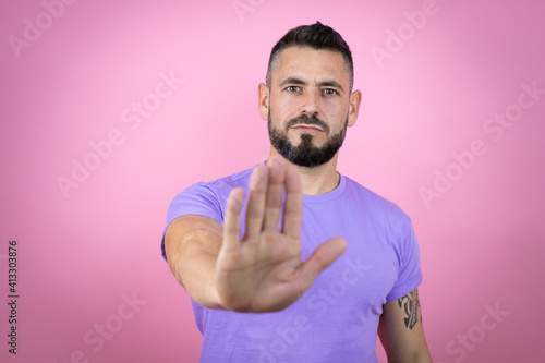 Young handsome man wearing casual t-shirt over pink background serious and doing stop sing with palm of the hand. © Irene