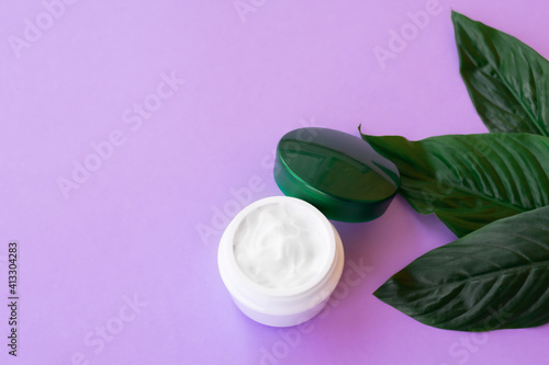 Soft white cream in jar with bright green leaves on purple background with copy space. Spa treatment and procedure at home.
