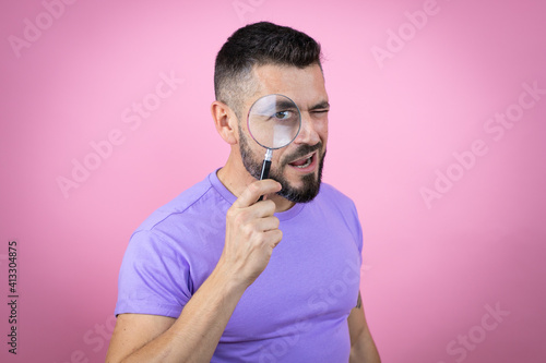 Young handsome man wearing casual t-shirt over pink background surprised looking through a magnifying glass