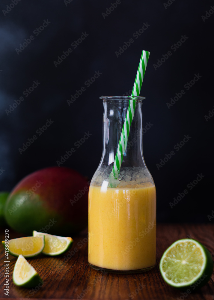 Fototapeta premium A fresh mango and lime smoothie in a glass milk bottle shape jar, with lime segments and mango fruits on table surface. Copy space available with dark background and wood table.
