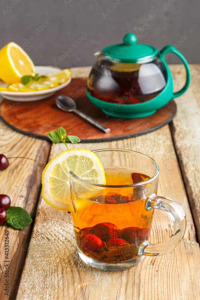 black tea in a glass cup with mint cherries and lemon on a wooden table and a teapot and lemon in a plate and mint leaves. black tea in a glass cup with mint cherries and lemon on a wooden table and a teapot and lemon in a plate and mint leaves.