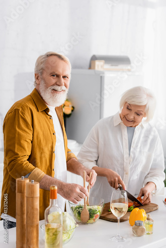 Senior man smiling at camera while mixing salad beside wife and wine on kitchen table