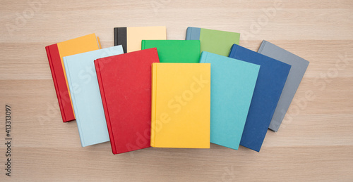 Top down view of a pile of bright colorful books centered on a light wood desk.