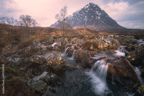 Moody, dramatic landscape of the iconic Buachaille Etiv Mor and River Coupall waterfall at Glencoe in the Scottish Highlands at sunrise or sunset.