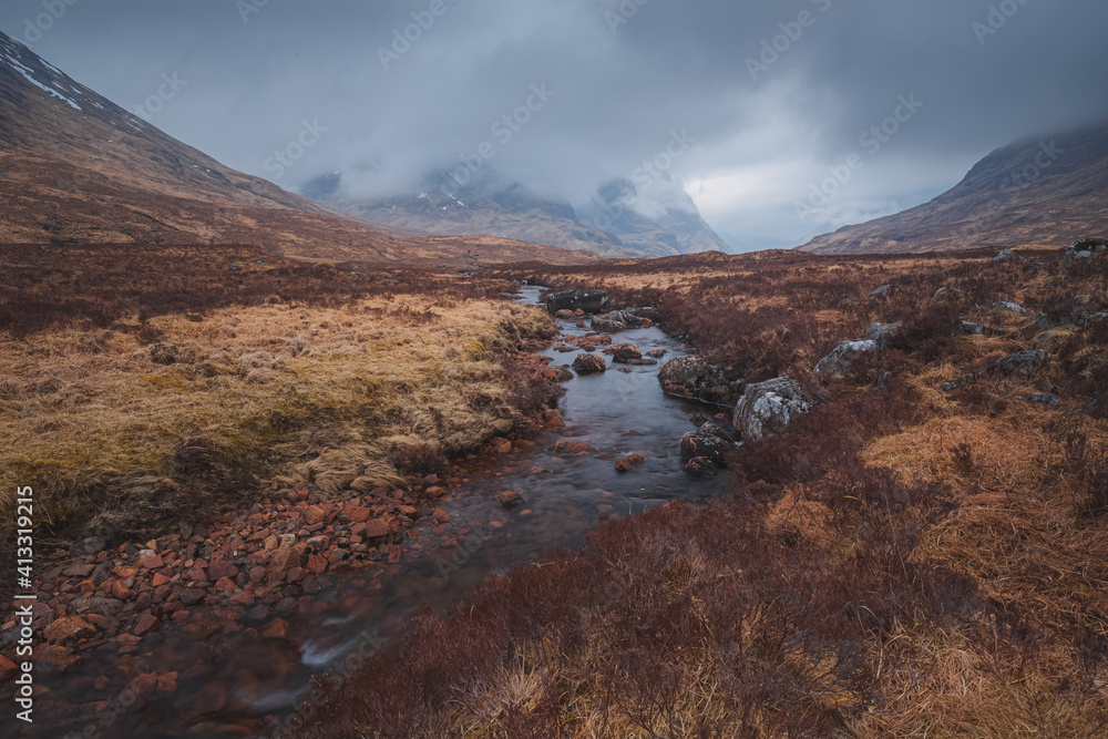 Dark and moody landscape of Glencoe in the Scottish Highlands with a foreground stream towards the ridges of Beinn Fhada, Gearr Aonach and Aonach Dubh (Three Sisters of Glencoe).