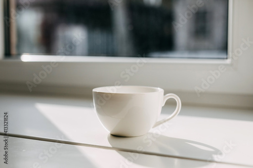 Blank white cup of coffee or tea on windowsill in sunlight.