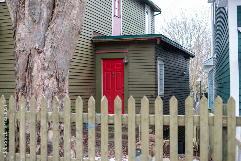 The side porch of a green building with a bright red metal door. A vintage small wooden window is on the side. A country style wooden picket fence and an old large oak tree are in the front yard. The side porch of a green building with a bright red metal door. A vintage small wooden window is on the side. A country style wooden picket fence and an old large oak tree are in the front yard.