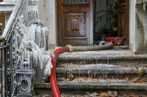 A cracked fire hose leaks water creating ice and icicles around stairs and fencing from a burning house. Soot is all around. photo