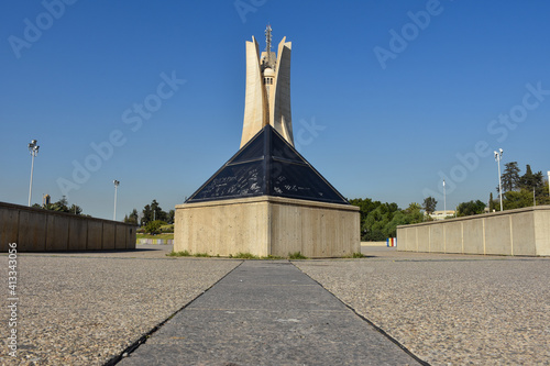 Low-angle perspective of the iconic Maqam Echahid (Martyrs' Memorial) and a dark glass pyramid in Algiers, Algeria, under a clear blue sky.