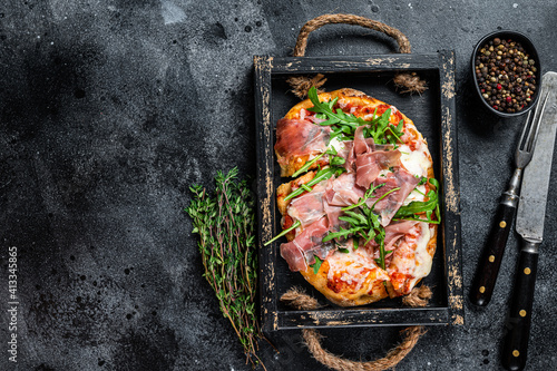 Italian Pizza with prosciutto parma ham, arugula salad and cheese in a rustic wooden tray. Black background. Top view. Copy space photo
