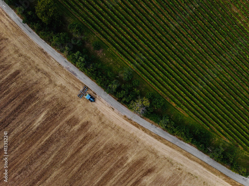 Aerial view of field in which tractor plows.Blue tillage equipment and blue tractor are clearly visible on brown background of earth.Road divides scape in two halves with green color in other one.