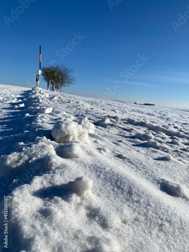 Zugeschneite Landstraße, Profil von Autoreifen zeichnen sich im Schnee ab. Straßenleitpfosten stehen am Rand, ein gelb schwarzer Schneeleitstab steht neben einem Pfosten. Im Horizont ein Baum