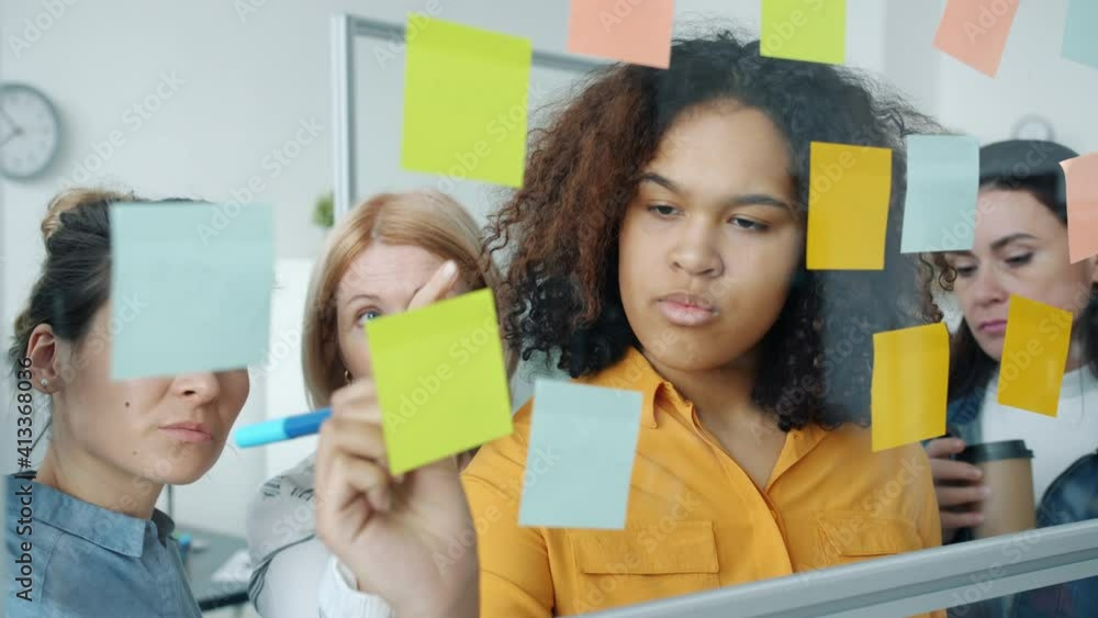 Group of women are talking and writing on colorful sticky notes working with glassboard in office together. Brainstorming and creative work concept. Group of women are talking and writing on colorful sticky notes working with glassboard in office together. Brainstorming and creative work concept.