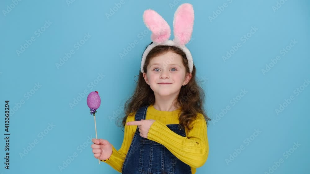 Positive cute little kid girl wears pink hare ears and jumper, points small colored eggs grow, makes holiday decorations, models over blue studio background. Look at this decorated Easter eggs! Positive cute little kid girl wears pink hare ears and jumper, points small colored eggs grow, makes holiday decorations, models over blue studio background. Look at this decorated Easter eggs!