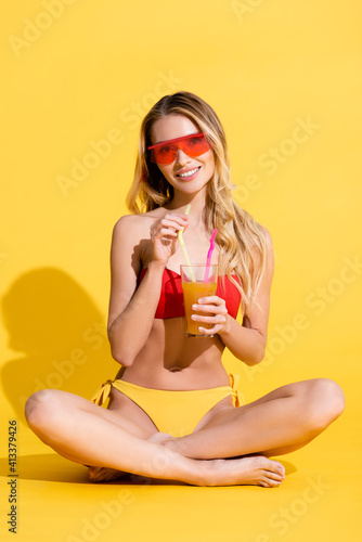 happy woman in swimsuit and sunglasses holding fresh cocktail while sitting with crossed legs on yellow.
