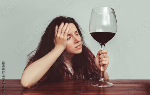 A young caucasian girl in a plaid shirt with tousled hair sits at a wooden table and holds a large glass of red wine.Concept of alcohol abuse, headache, alcoholism, hangover, loneliness and depression photo