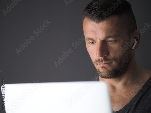 Man wearing gray t-shirt and wireless earphones sitting at table in home and working at laptop.