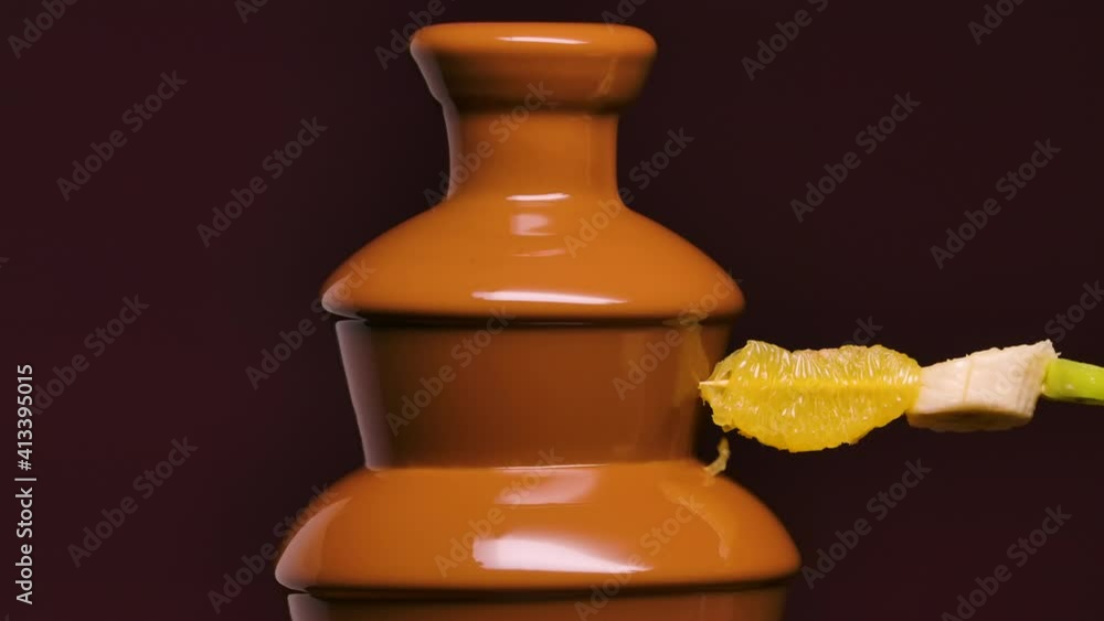 Melted milk chocolate flows in chocolate fountain. Ripe juicy fruit on skewer is dipped in sweet melted chocolate. Fondue. Dessert. Gourmet food. Party and celebration. Close up. Slow motion. Melted milk chocolate flows in chocolate fountain. Ripe juicy fruit on skewer is dipped in sweet melted chocolate. Fondue. Dessert. Gourmet food. Party and celebration. Close up. Slow motion.
