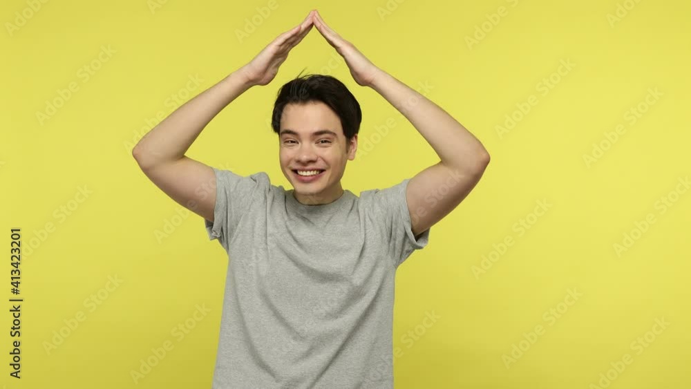 Happy smiling young man in casual gray t-shirt raising hands up making roof, concept of his own safety home, insurance agency, security service. Indoor studio shot isolated on yellow background Happy smiling young man in casual gray t-shirt raising hands up making roof, concept of his own safety home, insurance agency, security service. Indoor studio shot isolated on yellow background