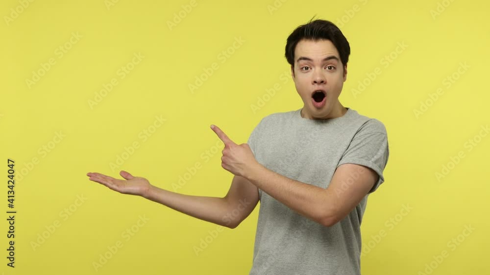 Attention at advertise! Surprised young man in gray t-shirt pointing to side, showing copy space for shocking news, final sale, crazy commercial text. Indoor studio shot isolated on yellow background Attention at advertise! Surprised young man in gray t-shirt pointing to side, showing copy space for shocking news, final sale, crazy commercial text. Indoor studio shot isolated on yellow background