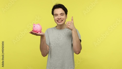 Cheerful positive young man in casual style gray t-shirt pointing at piggy bank and showing thumbs up, satisfied with money savings, deposit concept. Indoor studio shot isolated on yellow background photo
