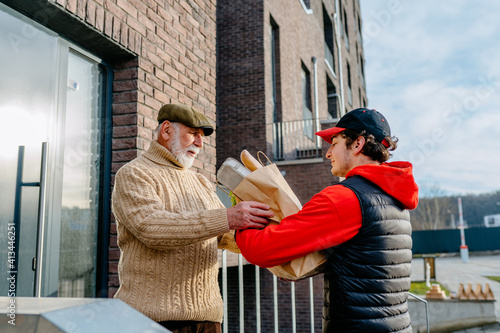 Rear of Caucasian young man from delivery service in red hat bringing packet with food to old man and giving it outdoors. Side view on female courier giving grocery deliver to grandfather.