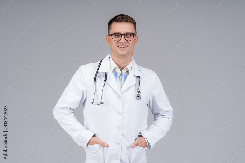 Practititoner in white scrubs put hands in pockets. Medic in glasses with phonendoscope on light background. Closeup. Practititoner in white scrubs put hands in pockets. Medic in glasses with phonendoscope on light background. Closeup.