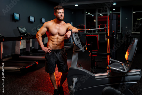 Athletic man without shirt poses to the camera with hand on waist. Dark light in gym. Modern interior. Closeup.