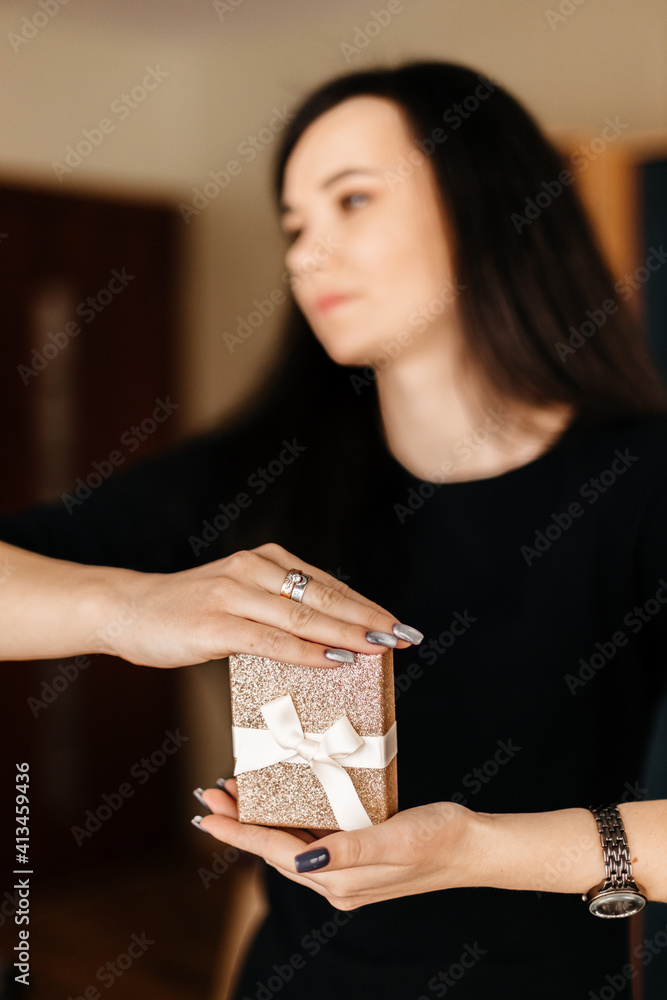 A beautiful brunette girl with long black hair in a black dress holds a golden gift box with a white ribbon bow A beautiful brunette girl with long black hair in a black dress holds a golden gift box with a white ribbon bow