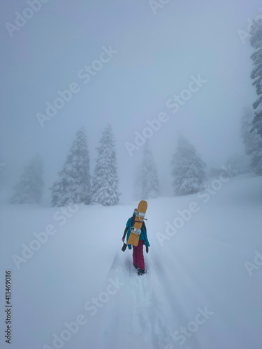 VERTICAL: Young female snowboarder hikes up a hill covered in fresh powder snow.