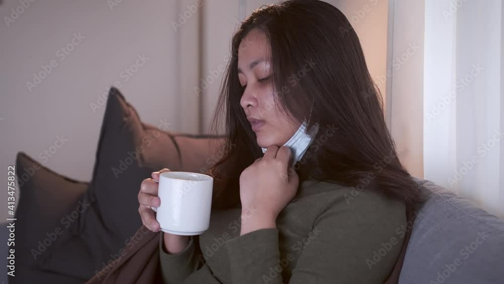 Sick Asian woman drinking lukewarm water in times of suffering has chilling black eyelids all over her body due to a cold and uses masks and blankets to warm and quarantine herself Sick Asian woman drinking lukewarm water in times of suffering has chilling black eyelids all over her body due to a cold and uses masks and blankets to warm and quarantine herself