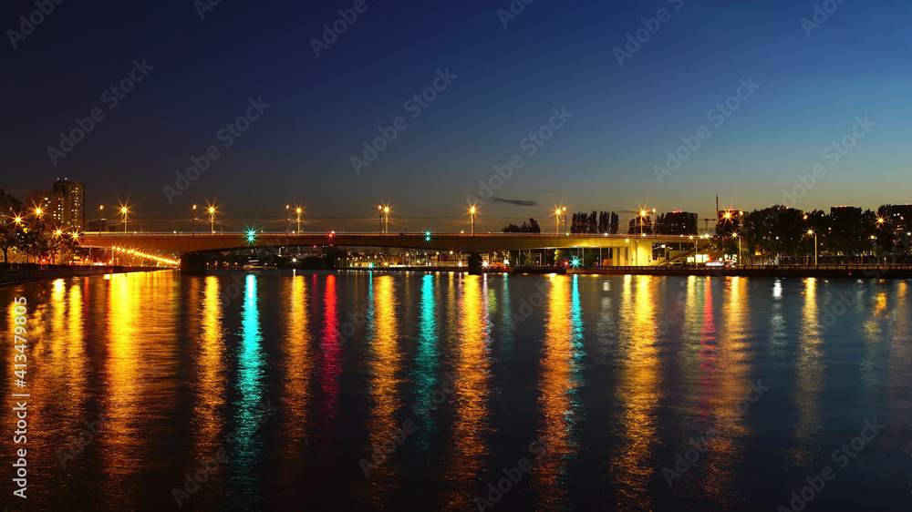 Bridge over the river at sunset with the reflection of city lighting in the water. Water surface in the twilight of the city.