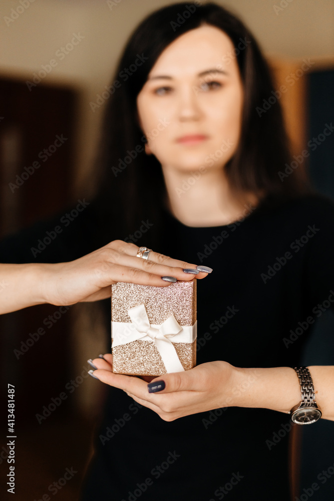 A beautiful brunette girl with long black hair in a black dress holds a golden gift box with a white ribbon bow A beautiful brunette girl with long black hair in a black dress holds a golden gift box with a white ribbon bow