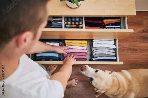 Organizing and cleaning home. Man preparing orderly folded t-shirts in drawer with his curious dog. photo