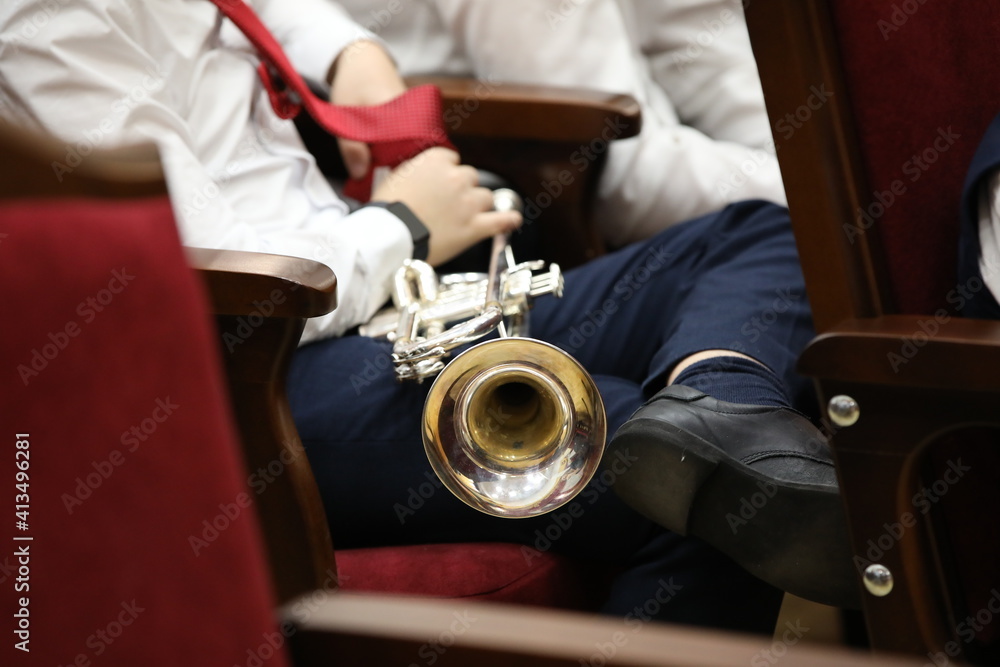A musical trumpet lies on the lap of a young musician in a white shirt and red tie sitting in a chair in the hall leg on leg during a concert.Background image selective focus