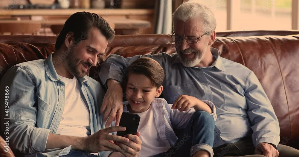 Smiling young man showing funny video on smartphone to laughing small boy son and sincere elderly mature father, relaxing together on comfortable couch, loving different generations family pastime. Smiling young man showing funny video on smartphone to laughing small boy son and sincere elderly mature father, relaxing together on comfortable couch, loving different generations family pastime.