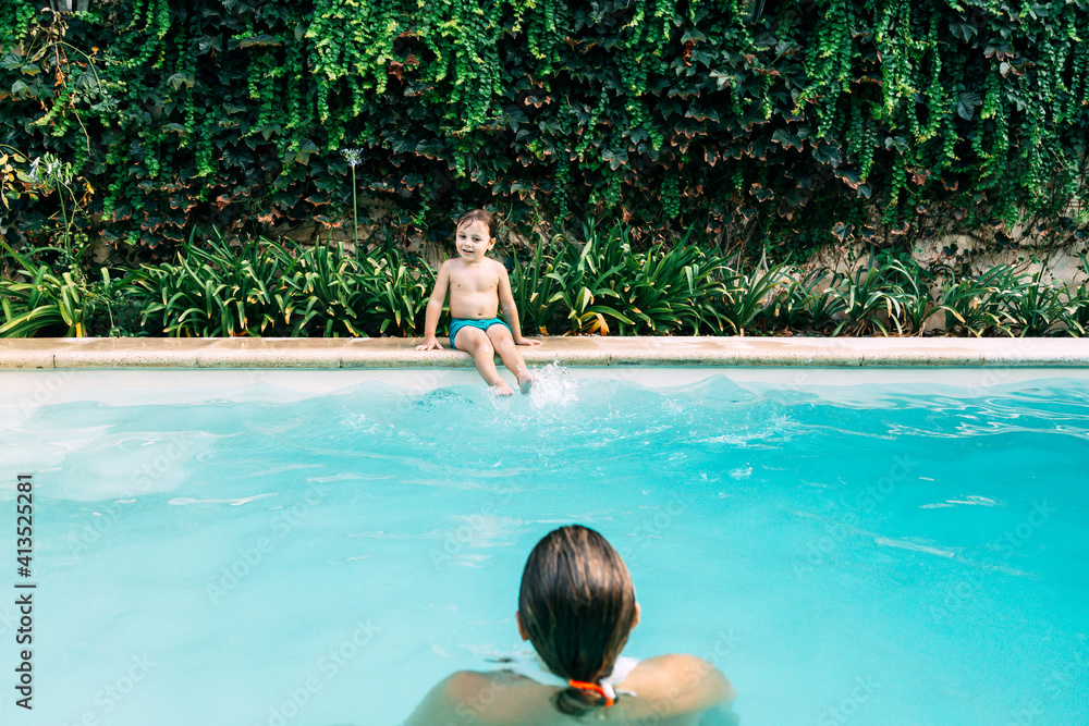 Little boy sitting in the border of swimming pool looking his mother into the pool.