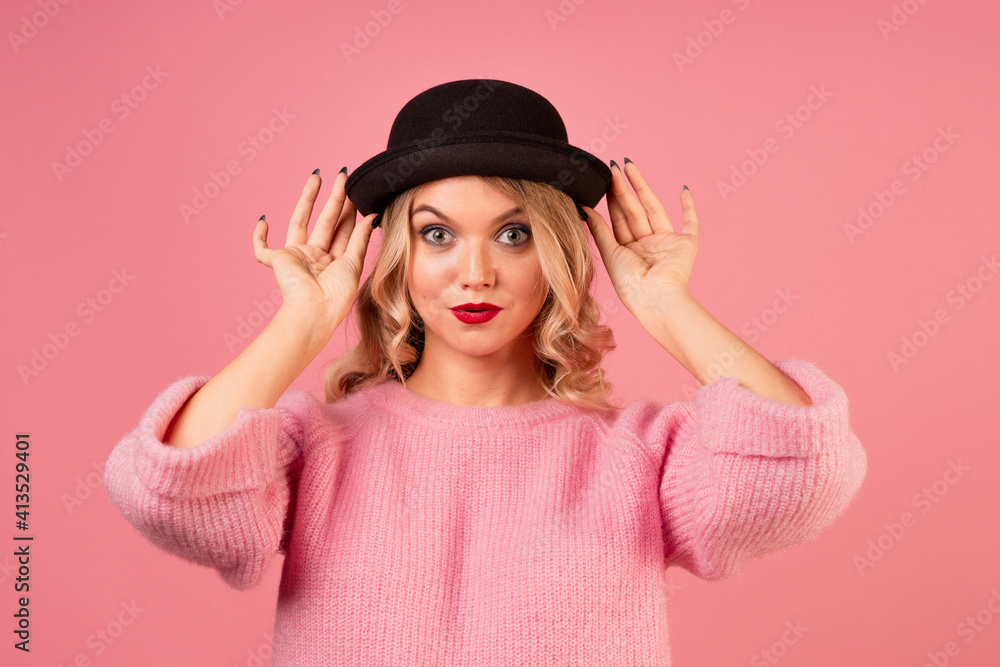 A young, attractive woman in a warm pink sweater and a black bowler hat on a pink background, blonde with curly hair and red lipstick A young, attractive woman in a warm pink sweater and a black bowler hat on a pink background, blonde with curly hair and red lipstick