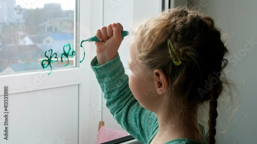 Drawing St. Patrick's Day Child painting green three-leaved shamrocks indoor, festive home decoration, quarantine family leisure. Little girl draws clover on window glass. Stay home concept New normal