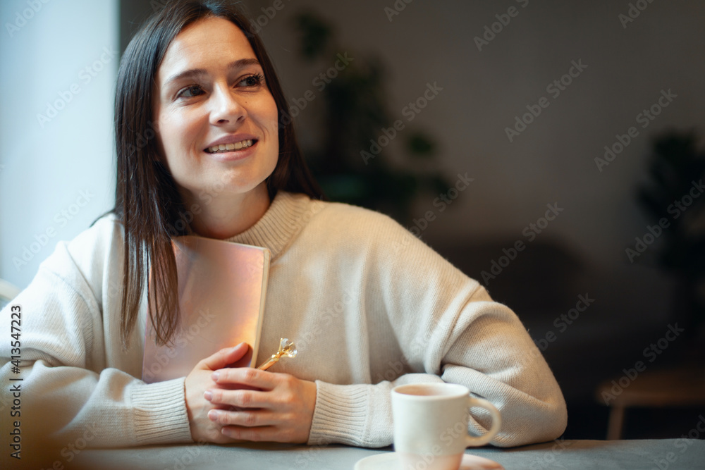 Inspired young woman with a smile looking through window and holding coral colored diary 2021 Inspired young woman with a smile looking through window and holding coral colored diary 2021